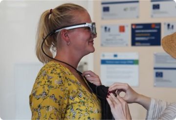 Mladá žena se světlými vlasy stojí bokem k nám. Jiná žena, která sotjí mimo záběr a vidíme jen její ruce, ji pomáhá nadadit si eye-trackingové brýle.