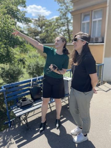 Mladá žena s tmavými vlasy v zeleném tričku ukazuje směr druhé ženě, stojící vedle ní. Druhá žena má nasazené eye-trackingové brýle.