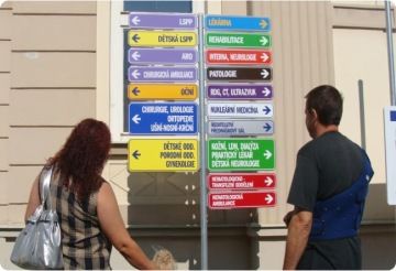 Žena s dítětem a muž s rukou v dlaze stojí zády k nám a dívají se na velmi barevný rozcestník se spoustou šipek